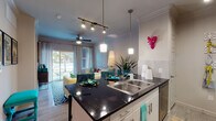 Located at 6600 McKinney Ranch Pkwy in McKinney, Texas, this single-floor residence offers a thoughtfully designed open-concept layout that seamlessly connects the kitchen, dining, and living areas. The kitchen is a standout feature, boasting dark granite countertops, a double-basin stainless steel sink, crisp white cabinetry, stainless steel appliances, and stylish pendant lighting paired with a track lighting system. Crown molding adds a refined architectural touch throughout the space. The living area is bright and welcoming, with a ceiling fan, sliding glass doors that invite natural light, and an outdoor patio view. Wood-style flooring flows consistently throughout the unit, adding warmth and continuity. The bedroom is a comfortable retreat, framed by crown molding, large windows dressed with teal drapes, and a ceiling fan. It connects directly to a private bathroom corridor that features a vanity with dark countertops and cabinetry, ample storage, and access to a spacious walk-in closet. The overall ambiance of the space is modern and inviting, with clean lines, neutral tones, and contemporary finishes that create a polished and functional living environment.