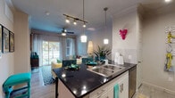 Located at 6600 McKinney Ranch Pkwy in McKinney, Texas, this single-floor residence offers a thoughtfully designed open-concept layout that seamlessly connects the kitchen, dining, and living areas. The kitchen is a standout feature, boasting dark granite countertops, a double-basin stainless steel sink, crisp white cabinetry, stainless steel appliances, and stylish pendant lighting paired with a track lighting system. Crown molding adds a refined architectural touch throughout the space. The living area is bright and welcoming, with a ceiling fan, sliding glass doors that invite natural light, and an outdoor patio view. Wood-style flooring flows consistently throughout the unit, adding warmth and continuity. The bedroom is a comfortable retreat, framed by crown molding, large windows dressed with teal drapes, and a ceiling fan. It connects directly to a private bathroom corridor that features a vanity with dark countertops and cabinetry, ample storage, and access to a spacious walk-in closet. The overall ambiance of the space is modern and inviting, with clean lines, neutral tones, and contemporary finishes that create a polished and functional living environment.