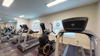 Located at Parkway Village in Grove City, Ohio, this well-equipped fitness center offers a comprehensive workout environment on a single floor. The space is organized into two distinct functional zones that together accommodate a full range of exercise routines.

The cardio area is lined with multiple treadmills, an elliptical trainer, a stationary bike, and a rowing machine, all positioned along walls featuring arched and standard windows fitted with horizontal blinds. Natural light filters in through these windows, creating a bright and welcoming atmosphere. A large wall mirror expands the sense of space and allows users to monitor their form during workouts.

The strength training section is equally well-appointed, housing a multi-station cable machine, an adjustable weight bench, and a tiered dumbbell rack stocked with a range of free weights. A wall-mounted flat-screen television provides entertainment during sessions, and full-length mirrors along one wall enhance both visibility and spatial openness.

Patterned modular carpet tiles in muted tones of grey and purple cover the floors throughout, providing cushioning and a polished aesthetic. The textured ceilings are illuminated by recessed fluorescent panel lighting. Additional amenities include stability balls, a hand sanitizer dispenser, and a convenient service window. The overall ambiance is clean, functional, and well-maintained, making it a practical fitness resource for residents.