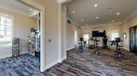 This single-floor community amenity facility offers a thoughtfully designed mix of fitness and leisure spaces, finished with consistent patterned carpet tile flooring and neutral warm-gray walls throughout. The fitness area is equipped with stationary bikes, treadmills, and a cable machine, complemented by wall-mounted mirrors and a large flat-screen TV. Recessed lighting and natural light from multiple windows create a bright, energetic environment. Adjacent to the fitness zone, a dedicated game room centers around a full-length shuffleboard table and features another large wall-mounted TV, with seating for spectators. A standout lounge area showcases a dramatic floor-to-ceiling stacked stone fireplace flanked by stylish lounge chairs with accent pillows, offering a warm and inviting retreat. Black-framed French doors and expansive windows allow ample natural light throughout. A separate seating nook with a glass-top table and modern blue chairs sits beneath large arched windows with outdoor views. A cozy media lounge provides additional seating and a mounted TV. Rounding out the amenities, a conveniently located snack kiosk with a refrigerated beverage case and packaged goods adds everyday convenience to the space.