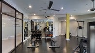This fitness center offers a thoughtfully designed workout space spread across a single floor. The room features dark, durable flooring that contrasts cleanly with the light gray walls and white trim, creating a sleek, modern aesthetic. Yellow accent columns add a bold visual element throughout the space, providing both structural support and a vibrant pop of color.

The free weight area is well-stocked with a full rack of dumbbells and a selection of medicine balls, complemented by adjustable workout benches positioned for versatile training. Large floor-to-ceiling mirrors line one wall, enhancing the sense of space while allowing users to monitor their form. The cardio and strength equipment section is visible toward the rear of the room, offering additional training options.

Overhead, two ceiling fans ensure consistent air circulation, supplemented by recessed lighting that provides bright, even illumination throughout the space. The layout is open and spacious, allowing for comfortable movement between equipment. A door toward the back suggests access to additional utility or storage areas, adding to the overall functionality of this well-appointed fitness facility.