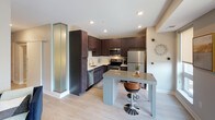 This single-floor space showcases a cohesive modern design with light wood flooring and a neutral color palette carried throughout. The kitchen is a standout feature, equipped with rich dark-toned shaker-style cabinetry extending from floor to ceiling, offering generous storage. A white quartz countertop contrasts elegantly against the cabinetry, complemented by a white subway tile backsplash. Stainless steel appliances, including a dishwasher, range, microwave, and refrigerator, add a contemporary finish. A central island with a gray quartz surface and bar seating provides additional prep space and casual dining. Frosted glass architectural columns flank the kitchen, adding a distinctive design element.

The living area is bright and welcoming, featuring large windows fitted with roller shades that allow natural light while maintaining privacy. A sliding glass door provides direct access to a balcony with outdoor views. The bathroom mirrors the home's design language, featuring dark shaker-style cabinetry, a white countertop vanity, a framed mirror, and a well-lit vanity fixture above. The overall ambiance is clean, contemporary, and thoughtfully designed, with consistent finishes tying each space together seamlessly.