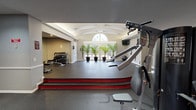 The Canter Chase fitness center is a thoughtfully designed, single-floor facility that offers a comprehensive workout environment across multiple interconnected zones. The space is architecturally distinguished by a vaulted, beam-accented ceiling in the studio area, which features a dramatic arched window that floods the room with natural light and views of the surrounding outdoor greenery. Four large potted palm trees line the window wall, adding a refreshing, natural ambiance to the space. Rubber flooring throughout provides durability and comfort underfoot, with a raised platform area in the studio suitable for group exercise or stretching. The cardio zone is equipped with treadmills, elliptical machines, and upright bikes, positioned near a wide picture window with outdoor views and ceiling fans overhead for added comfort. A wall-mounted television is conveniently accessible from this area. The strength training section includes a multi-station cable machine, a full dumbbell rack with accompanying benches, and mirrored walls that enhance the sense of space. Indirect cove lighting with a curved ceiling detail creates a warm, polished atmosphere in the main gym area. TRX suspension straps, a stability ball, and additional functional training equipment round out the offerings, making this a fully equipped community fitness facility.