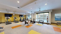Located at 3808 South West Temple Street in South Salt Lake, Utah, this fitness center is a comprehensively outfitted exercise facility designed to support a wide range of workout routines. The space is organized across a single floor and divided into distinct functional zones, each thoughtfully arranged to maximize usability and flow.

The cardio area features multiple treadmills, elliptical trainers, and stationary cycling bikes, positioned to take advantage of large windows that invite natural light and provide views of the surrounding outdoor areas. A wall-mounted television and ceiling fans add comfort and convenience to the cardio zone.

Adjacent areas are dedicated to strength training, including a multi-station cable machine, a weight bench with free weights, and a full dumbbell rack. Mirrored walls in the strength section enhance the sense of space and allow users to monitor their form during exercise.

A separate cycling studio accommodates several Spinning bikes alongside stability balls, creating a dedicated space for group or individual cycling workouts. A water fountain and storage shelving are conveniently situated nearby.

Throughout the facility, the neutral carpet tile flooring is accented with bold orange and yellow geometric inlays, adding a modern, energetic aesthetic. Recessed lighting and ceiling fans maintain a bright and comfortable environment. The overall design is clean, functional, and well-maintained, offering a complete fitness experience.