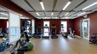 This fitness center is designed to support a comprehensive workout experience within a single, open-plan floor. The space immediately draws attention with its striking deep crimson walls, which create an energetic and motivating atmosphere. Overhead, a grid-style ceiling fitted with long fluorescent light panels ensures the entire space is evenly and brightly illuminated, complementing the natural light that streams in through multiple windows along the back wall.

The flooring consists of smooth hardwood-style planks, offering a clean and durable surface suitable for a wide range of exercises. A row of treadmills with individual display screens is centrally positioned, forming the core of the cardio zone. Flanking them are additional cardio machines, including an elliptical and a stationary bike, providing varied aerobic training options.

Along the left wall, large mirrors amplify the sense of space and allow users to monitor their form. A well-stocked free weight and accessory station is also visible, featuring medicine balls in various weights, a stability ball, and other training accessories neatly arranged for easy access. A wall-mounted television screen adds an entertainment element to cardio sessions. The overall ambiance is purposeful and energizing, making this fitness center a practical and fully functional facility.