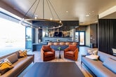 This contemporary amenity lounge is thoughtfully designed across a single floor, offering a seamless flow between distinct social and entertainment zones. The main lounge area features warm wood-look tile flooring throughout, complemented by an eclectic mix of upholstered seating including rich burnt-orange velvet armchairs, light gray sectional sofas, and structured accent chairs. A dramatic floor-to-ceiling teal textured wall panel serves as a bold focal point, anchoring a café-style seating area furnished with round tables and sleek bistro chairs. Recessed lighting and pendant fixtures illuminate the space, while a wall-mounted flat-screen TV adds a media entertainment element. A full bar with a dark stone countertop and leather-upholstered bar stools lines one wall, accompanied by vertical dark wood slat paneling that adds depth and warmth. A separate game room features a professional billiards table beneath a striking geometric brass chandelier, with large floor-to-ceiling windows offering direct views of an outdoor pool area. Sliding dark fluted wood panel partitions allow for flexible spatial configurations. Natural stone accent walls and lush indoor greenery complete the refined, resort-inspired ambiance of this well-appointed communal space.