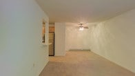 This single-floor residence offers a clean, functional layout designed for comfortable everyday living. The living area is generously proportioned, featuring neutral-toned carpet flooring that adds warmth underfoot. A ceiling fan with an integrated light fixture serves as the focal point of the room, providing both airflow and ambient lighting. A baseboard heating unit runs along the far wall, ensuring year-round comfort. The living space flows naturally toward the kitchen, which is partially separated by a structural column and a raised breakfast bar — ideal for casual dining or entertaining. The kitchen is equipped with wood-toned cabinetry offering ample storage, a full-size refrigerator, and tile flooring that contrasts neatly with the carpeted living area. The overall palette is light and neutral throughout, with crisp white walls that lend an airy, open feel to the interior. The layout is straightforward and adaptable, making efficient use of the available space. The combination of functional features and an open-concept connection between the kitchen and living area creates a cohesive and practical living environment well-suited for a variety of lifestyles.