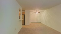 This single-floor residence offers a clean, functional layout designed for comfortable everyday living. The living area is generously proportioned, featuring neutral-toned carpet flooring that adds warmth underfoot. A ceiling fan with an integrated light fixture serves as the focal point of the room, providing both airflow and ambient lighting. A baseboard heating unit runs along the far wall, ensuring year-round comfort. The living space flows naturally toward the kitchen, which is partially separated by a structural column and a raised breakfast bar — ideal for casual dining or entertaining. The kitchen is equipped with wood-toned cabinetry offering ample storage, a full-size refrigerator, and tile flooring that contrasts neatly with the carpeted living area. The overall palette is light and neutral throughout, with crisp white walls that lend an airy, open feel to the interior. The layout is straightforward and adaptable, making efficient use of the available space. The combination of functional features and an open-concept connection between the kitchen and living area creates a cohesive and practical living environment well-suited for a variety of lifestyles.