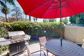 Located at 14731 Sherman Way in Los Angeles, CA, this single-level outdoor space offers a well-appointed patio area designed for comfortable outdoor living and entertaining. The patio is centered around a circular slatted dining table accompanied by multiple chairs and shaded by a large red market umbrella, providing ample cover from the Southern California sun. A full-size stainless steel gas BBQ grill is positioned nearby, making the space well-suited for outdoor cooking and gatherings. The patio is enclosed by a dense, lush ivy-covered hedge that lines the perimeter, offering a sense of privacy while adding a vibrant, garden-like atmosphere. Flowering vines with purple blooms further enhance the greenery, creating a visually rich border. Mature palm and shade trees are visible beyond the hedge, contributing to the quintessential Los Angeles landscape. The building exterior features a stucco finish, with raised planters filled with well-maintained shrubs adding additional greenery along the structure's base. A gated entry provides access to the surrounding area, while exterior lighting fixtures and a staircase with metal railings are also visible, reflecting a practical and clean architectural design. The space benefits from abundant natural sunlight throughout the day.