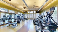 This fitness center offers a generously proportioned, single-level workout space designed to accommodate a wide range of exercise needs. The room features notably high ceilings with exposed ductwork and multiple ceiling fans suspended throughout, promoting airflow and contributing to an open, airy atmosphere. Large windows fitted with blinds line the walls, allowing ample natural light to fill the space while providing views of the surrounding exterior.

The flooring consists of durable, multi-toned resilient material in yellow and brown tones, clearly delineating workout zones throughout the room. Full-length mirrors are mounted along one wall, enhancing the sense of depth and allowing users to monitor their form. The walls are painted in warm yellow and green tones, creating an energetic yet welcoming environment.

The cardio zone is well-stocked with treadmills, elliptical trainers, stair climbers, and stationary bikes, many equipped with individual monitors. The strength training area includes cable machines, a functional trainer, selectorized weight machines, adjustable benches, and a comprehensive free weight section with dumbbells and kettlebells. Suspended linear lighting supplements the natural light, ensuring consistent illumination throughout the entire facility.