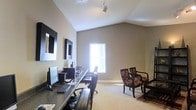 Located at 3851 Cape Landing Drive in Myrtle Beach, South Carolina, this single-story property offers a well-appointed and functional layout designed for comfort and practicality. The space features a dedicated office or study area that immediately stands out for its vaulted ceilings, which lend a sense of openness and height to the room. Neutral-toned walls in a warm greige palette create a calm, focused atmosphere, while carpeted flooring adds a comfortable feel underfoot. The office is equipped with a long, dark-toned desk surface running along one wall, accommodating multiple monitors and workstations, making it well-suited for productivity. Track lighting on the ceiling provides targeted illumination, complementing the natural light that filters in through a centrally placed window. A seating area within the office features patterned upholstered armchairs and a side table, offering a comfortable spot for reading or informal meetings. Decorative framed mirrors on the wall add visual depth and a polished finishing touch. The textured ceiling detail is consistent throughout, adding a classic architectural element to the overall design of the home.