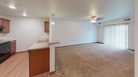 This single-floor residential unit offers a practical and comfortable layout designed for everyday living. The open-concept kitchen and living area form the heart of the space, where warm medium-toned wood cabinetry pairs with a peninsula countertop that doubles as a breakfast bar. Black appliances, including a refrigerator and electric range, are set beneath a ventilation hood, while recessed and pendant lighting illuminate the space. The kitchen transitions seamlessly to the living area, which features soft wall-to-wall carpeting and a ceiling fan with light fixture. Light wood-tone laminate flooring runs through the entry and kitchen zone, creating a clear distinction between functional and living spaces. A dedicated in-unit laundry room houses a full-size top-load washer and dryer with wire shelving above for added storage. The bedroom is a quiet, carpeted room with a window fitted with horizontal blinds that allow for natural light and privacy. The bathroom is clean and functional, featuring a single-sink vanity with medium wood cabinetry, a large mirror, tile flooring, and a full tub-and-shower combination. Throughout the unit, white walls and wood-toned trim details contribute to a cohesive, understated aesthetic.