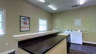 This single-story laundry facility is designed with practicality and efficiency in mind. The room features neutral sage-green walls and durable dark-stained countertops that provide generous folding and sorting surface area. A row of washers and dryers lines the back wall, offering multiple machines to accommodate high-volume use. A built-in utility sink is conveniently positioned near the machines, making it easy to pre-treat garments or handle hand-wash items. Overhead fluorescent lighting ensures the space is well-lit throughout, while a window along the back wall introduces natural light and a view of surrounding greenery, adding a refreshing touch to the utilitarian environment. The dark hardwood-style flooring contrasts neatly with the lighter walls and cabinetry. An informational bulletin board on the wall adds a community-oriented element to the space. Overall, the facility is clean, organized, and thoughtfully equipped to serve as a fully functional shared laundry room.