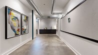 Located at 423 E 36th Street in Charlotte, NC, this single-floor creative space blends industrial design with vibrant, curated interiors. Polished concrete floors run throughout, complementing exposed silver ductwork overhead and crisp white walls that give the space an open, loft-like character. Structural columns finished in deep teal blue serve as architectural accents, tying together the design palette.

The main corridor doubles as a gallery-style hallway, showcasing bold large-format portrait artwork in black frames. A sizable corkboard panel and additional wall-mounted display surfaces line the hallway, offering functional workspace alongside the artistic aesthetic. At one end, a sleek black kitchenette with a sink and bar-height seating provides a practical refreshment area set against a white tile backsplash.

The lounge area is a standout feature, furnished with a chartreuse sofa, a warm orange armchair, and a patterned geometric area rug in black and white. Whimsical bowler hat-shaped wall sconces illuminate a grid-patterned accent wall, creating a welcoming and eclectic atmosphere. A floor lamp and curated wall art complete the vibe. A green screen wall visible from the open floor plan adds a versatile, production-ready element to the space. Throughout, the design strikes a balance between functional utility and creative energy, making it well-suited for professional, artistic, or collaborative use.