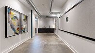 Located at 423 E 36th Street in Charlotte, NC, this single-floor creative space blends industrial design with vibrant, curated interiors. Polished concrete floors run throughout, complementing exposed silver ductwork overhead and crisp white walls that give the space an open, loft-like character. Structural columns finished in deep teal blue serve as architectural accents, tying together the design palette.

The main corridor doubles as a gallery-style hallway, showcasing bold large-format portrait artwork in black frames. A sizable corkboard panel and additional wall-mounted display surfaces line the hallway, offering functional workspace alongside the artistic aesthetic. At one end, a sleek black kitchenette with a sink and bar-height seating provides a practical refreshment area set against a white tile backsplash.

The lounge area is a standout feature, furnished with a chartreuse sofa, a warm orange armchair, and a patterned geometric area rug in black and white. Whimsical bowler hat-shaped wall sconces illuminate a grid-patterned accent wall, creating a welcoming and eclectic atmosphere. A floor lamp and curated wall art complete the vibe. A green screen wall visible from the open floor plan adds a versatile, production-ready element to the space. Throughout, the design strikes a balance between functional utility and creative energy, making it well-suited for professional, artistic, or collaborative use.