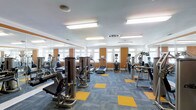 This fitness center offers a spacious, single-floor layout designed to accommodate a comprehensive workout experience. The room is filled with an extensive selection of equipment, including treadmills, ellipticals, stair climbers, and a variety of strength-training machines, all arranged to allow comfortable movement between stations. Large windows line the walls, inviting abundant natural light and providing views of the surrounding exterior, contributing to an open and energizing atmosphere. The ceiling is finished in white with recessed fluorescent lighting panels that ensure the space remains consistently well-lit throughout. A full-length mirror runs along one wall, enhancing the sense of depth and allowing users to monitor their form during workouts. The flooring features a distinctive blue and yellow modular carpet tile pattern, adding a dynamic visual element to the space. Orange accent panels on the walls complement the color scheme, giving the fitness center a cohesive and modern aesthetic. The overall ambiance is clean, organized, and purpose-driven, making it a functional and inviting environment suited for a wide range of fitness activities.