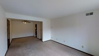 This single-floor space features a clean, functional layout with a straightforward design. The main living area is finished with wall-to-wall neutral-toned carpet and smooth white walls, creating a blank canvas that is easy to personalize. Dark baseboards provide a subtle contrast along the perimeter of the room. A wide doorway connects the main living area to an adjacent room, enhancing the sense of openness and flow between spaces. The adjoining room is equipped with a ceiling fan with integrated lighting, offering both air circulation and ambient illumination. Through the doorway, additional built-in storage or cabinetry is partially visible, suggesting practical utility in the connected space. Electrical outlets are positioned at regular intervals along the walls, accommodating a variety of furniture arrangements. The overall ambiance is understated and neutral, with consistent finishes throughout. The absence of decorative elements keeps the focus on the functional aspects of the layout, making it a versatile space well-suited to a range of uses and personal styles.