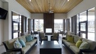This covered lanai is a thoughtfully designed outdoor living space that seamlessly blends indoor comfort with an open-air atmosphere. The ceiling is finished with rich, warm-toned wood planking, lending a natural elegance that contrasts beautifully against the crisp white walls. Recessed lighting and a modern ceiling fan ensure the space remains comfortable and well-lit throughout the day and into the evening.

The lanai is framed by large black-framed glass panels and doors that create a striking architectural boundary between the outdoor lounge and the interior space beyond, allowing for clear sightlines and an abundance of natural light. Dark vertical accent columns punctuate the openings, adding a bold, contemporary design element.

The seating area is furnished with dark wicker lounge chairs and sofas accented with neutral cushions and colorful throw pillows in shades of teal, green, and gold, creating an inviting and vibrant atmosphere. A wicker coffee table anchors the central seating arrangement, making it an ideal gathering spot.

Beyond the glass walls, views of the surrounding outdoor amenities — including what appears to be a pool area lined with palm trees — further enhance the resort-like ambiance of this well-appointed lanai space.