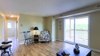 This single-floor space centers around a welcoming living area finished with warm-toned hardwood flooring that extends throughout, lending a cohesive and clean aesthetic to the interior. The walls are painted in a soft, neutral yellow-green hue that complements the natural light flowing in from a large sliding glass door, which opens directly onto a balcony overlooking lush greenery. The balcony access serves as a standout feature, effectively blending indoor and outdoor living while providing pleasant views of the surrounding landscape.

The living area is thoughtfully laid out, offering ample room for seating arrangements and everyday use. Decorative wall plates add a subtle artistic touch above the sliding door, while a classic table lamp provides warm accent lighting. The textured ceiling adds a layer of architectural character to the space. Ventilation is supported by ceiling vents, ensuring year-round comfort. Neutral-toned baseboards and trim provide clean definition throughout the room. Overall, the space strikes a balance between casual comfort and functional design, making it an inviting and practical environment suited to a variety of living arrangements.