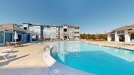 This outdoor amenity space in Macon, Georgia, is centered around a generously sized freeform swimming pool with vivid blue water, offering a resort-like atmosphere within a residential community setting. The pool area is framed by a broad concrete sundeck furnished with lounge chairs, outdoor seating sets, and shaded umbrella tables, providing ample space for relaxation and socializing under the Georgia sun.

A standout feature is the covered outdoor pavilion, finished in a coastal-inspired blue lap siding with crisp white trim. Ceiling fans overhead ensure comfort in warmer months, while a built-in bar counter and high-top seating create a functional and inviting gathering spot. Draping curtain panels lend a cabana-style aesthetic to the space.

The surrounding grounds include manicured open lawn areas and mature tree lines that provide a natural backdrop. A black iron perimeter fence defines the space while maintaining open sightlines to the wooded surroundings. The multi-story residential building, clad in blue siding with white-trimmed balconies and dormer accents, anchors the community visually and overlooks the pool area. The overall ambiance is bright, clean, and welcoming, suited for outdoor living throughout the seasons.