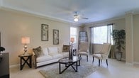 This 3-floor home offers a refined and comfortable living environment, highlighted by the inviting main living area visible in the space. The living room features light beige walls accented by crown molding, creating a polished architectural finish. Large windows dressed with blinds allow abundant natural light to fill the room, contributing to its airy, open atmosphere. Light-toned tile flooring runs throughout, complemented by a plush area rug that grounds the seating arrangement. The room is centered around a cream sofa adorned with textured throw pillows, paired with two elegant high-backed accent chairs in a warm neutral fabric. A glass-topped coffee table with a dark metal X-frame base adds a contemporary touch to the space. Decorative framed wall art featuring botanical motifs enhances the room's refined aesthetic. A ceiling fan with an integrated light fixture provides both function and subtle style. The overall ambiance of the home is calm, elegant, and well-balanced, blending classic architectural details with modern furnishings across its three floors to create a cohesive and welcoming living environment.