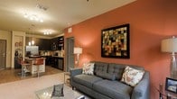 This single-floor space offers a thoughtfully designed open-concept layout that seamlessly connects the living area and kitchen, creating a cohesive and functional environment. The living room is anchored by a bold terracotta accent wall that adds warmth and character to the space, complemented by a contemporary tufted gray sofa adorned with decorative throw pillows. A striking large-format mosaic-style artwork serves as a focal point on the accent wall, adding a vibrant and artistic touch to the room. The space is illuminated by ambient lighting, including a classic table lamp that contributes to the inviting atmosphere. The kitchen, visible from the living area, features dark cabinetry with modern hardware, stainless steel appliances including a microwave and dishwasher, and a decorative tile backsplash that adds visual interest. Light hardwood-style flooring flows throughout, tying the kitchen and living areas together cohesively. A glass-top coffee table in the foreground enhances the modern aesthetic, while the overall color palette of warm terracotta, charcoal, and neutral tones creates a balanced and stylish ambiance throughout this well-appointed space.