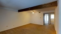 This single-story space offers a straightforward and functional layout designed for comfortable everyday living. The main living area is immediately notable for its striking exposed dark wood beam that spans the ceiling, adding a rustic architectural contrast to the otherwise clean, white walls. A ceiling fan with a light fixture is centrally positioned, providing both air circulation and ambient lighting to the room. The floors throughout are covered in wall-to-wall carpet in a warm, neutral tone, lending a cozy feel underfoot. The textured ceiling adds subtle visual interest while maintaining a classic residential aesthetic. Crisp white walls keep the space feeling bright and open, offering a neutral canvas ready for personalization. A doorway visible in the background leads to an additional area that benefits from natural light, suggesting a secondary room or entry point with exterior door access. Electrical outlets and HVAC vents are thoughtfully placed throughout, ensuring practical comfort. Overall, the space presents a clean, move-in ready environment with character details like the exposed beam that set it apart from more standard interiors.