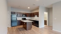 This single-floor residence is designed with a functional, open layout that connects its key living spaces seamlessly. The kitchen serves as a central highlight, outfitted with rich dark espresso cabinetry, granite-look countertops, and a full suite of black appliances including a refrigerator, range, microwave, and dishwasher. A substantial center island with a matching granite surface provides additional workspace and casual dining potential. Light wood-style flooring runs throughout, contributing to a clean, cohesive aesthetic.

A hallway leads from the kitchen toward the bedroom areas, with a large walk-in closet featuring wire shelving systems that maximize vertical and hanging storage. A second walk-in closet offers comparable storage capacity. The bathroom includes a dark wood vanity topped with granite, an undermount sink, and brushed nickel fixtures.

The living area is generously proportioned, featuring a ceiling fan, large windows that bring in natural light, and direct access to an exterior door. Neutral gray walls throughout the unit create a versatile backdrop. The overall design is modern and consistent, with attention to practical details and quality finishes across every room.
