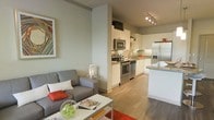 This single-floor space showcases a thoughtfully designed open-concept layout that integrates the living area and kitchen into one cohesive environment. The living area is anchored by a contemporary gray sofa accented with colorful throw pillows, a sleek glass-topped coffee table, and a vibrant piece of wall art, creating an inviting atmosphere. Warm hardwood-style flooring flows throughout, tying the spaces together visually.

The kitchen is well-appointed with stainless steel appliances, including a refrigerator with ice maker, a range, and an over-the-range microwave. Crisp white cabinetry extends to the upper walls, providing ample storage, while the granite-topped island serves as both a functional workspace and a casual dining surface. Under-cabinet lighting enhances the work areas, and track lighting overhead ensures the kitchen remains bright and practical.

A partial wall subtly separates the living and kitchen zones while maintaining the open feel of the space. The overall ambiance is clean, modern, and functional, with neutral tones complemented by strategic pops of color throughout.