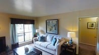 This single-floor space showcases a comfortable and thoughtfully designed living room as its centerpiece. The room features warm, neutral-toned walls that create an inviting ambiance, complemented by a textured ceiling that adds subtle visual interest. A large window dressed with dark valances and teal curtain panels allows natural light to filter in, brightening the space and offering a view to the outdoors.

The living room is anchored by a plush, light gray sofa adorned with teal and patterned throw pillows, adding pops of color that tie the room's palette together. A prominent framed abstract artwork in muted greens, blues, and grays serves as a focal point on the main wall, enhancing the room's aesthetic character. Two coordinating table lamps with ceramic teal bases flank the seating area, providing warm ambient lighting. Dark wood end tables and a matching coffee table ground the space with contrast against the lighter furnishings. A area rug defines the seating zone over the carpeted floor. The overall atmosphere is relaxed, cohesive, and welcoming, with decorative touches that reflect a carefully curated interior style.
