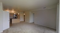 This single-floor residence offers a clean, open-concept layout designed for comfortable everyday living. The expansive living area is finished with plush wall-to-wall carpeting in a warm neutral tone, creating a soft and inviting atmosphere. Smooth walls painted in a crisp off-white palette lend the space a bright, airy feel, complemented by centrally placed ceiling vents that ensure consistent climate control throughout.

The kitchen is positioned adjacent to the living area and features light wood cabinetry that provides ample storage. Black appliances, including a built-in microwave and range, add a modern contrast to the cabinetry. A peninsula counter with pendant lighting overhead defines the kitchen space while maintaining its open connection to the living area, making it well-suited for both cooking and casual entertaining.

Several interior doors lead to additional rooms and closet space, including what appears to be a generously sized closet off the main living area. The layout flows intuitively, with clear sightlines across the open floor plan. Overall, the space presents a practical and well-organized design with consistent finishes, neutral décor, and functional features that create a welcoming and versatile environment.