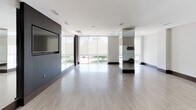 This single-floor fitness studio is designed with a clean, minimalist aesthetic that prioritizes open space and functionality. The main workout area is expansive, featuring light-toned hardwood flooring that extends throughout, creating a bright and welcoming environment suited for a variety of exercise disciplines.

A defining architectural feature is the floor-to-ceiling window wall along one side of the space, fitted with roller shades to manage natural light. These large-format windows offer views of the surrounding outdoor landscape and flood the interior with abundant daylight. Full-wall mirrors are strategically positioned throughout the studio, visually expanding the space while serving as practical tools for form and movement monitoring.

A dark wood-paneled accent wall houses a wall-mounted flat-screen television, providing a focal point for guided workouts or media display. Adjacent to the main floor, a glass-door enclosure offers a separate, more private zone for training or storage. Wall-mounted accessory holders, a stability ball, and additional fitness equipment are integrated throughout. Recessed ceiling lighting and ceiling-mounted speakers complete the polished, professional atmosphere of this well-appointed fitness space.
