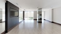 This single-floor fitness studio is designed with a clean, minimalist aesthetic that prioritizes open space and functionality. The main workout area is expansive, featuring light-toned hardwood flooring that extends throughout, creating a bright and welcoming environment suited for a variety of exercise disciplines.

A defining architectural feature is the floor-to-ceiling window wall along one side of the space, fitted with roller shades to manage natural light. These large-format windows offer views of the surrounding outdoor landscape and flood the interior with abundant daylight. Full-wall mirrors are strategically positioned throughout the studio, visually expanding the space while serving as practical tools for form and movement monitoring.

A dark wood-paneled accent wall houses a wall-mounted flat-screen television, providing a focal point for guided workouts or media display. Adjacent to the main floor, a glass-door enclosure offers a separate, more private zone for training or storage. Wall-mounted accessory holders, a stability ball, and additional fitness equipment are integrated throughout. Recessed ceiling lighting and ceiling-mounted speakers complete the polished, professional atmosphere of this well-appointed fitness space.