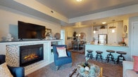 This single-floor residence showcases an open-concept design that seamlessly connects the living, dining, and kitchen areas into one cohesive and functional space. The living area is anchored by a striking mosaic-tiled fireplace, which serves as a focal point, complemented by plush carpeting and a tray ceiling detail that adds architectural depth overhead. A large flat-screen television is mounted above the fireplace, enhancing the room's entertainment appeal.

Adjacent to the living space, the dining area accommodates a full dining table and chairs, positioned beneath a chandelier that adds a warm, inviting glow. The open sightlines between the spaces create a sense of generous scale and ease of flow throughout.

The kitchen features white cabinetry, granite-style countertops, stainless steel appliances, and a peninsula with bar-height seating — ideal for casual dining or entertaining. Pendant lighting above the peninsula adds a polished finishing touch. A textured ceiling and consistent neutral tones throughout contribute to a cohesive, contemporary aesthetic. Overall, the space balances comfort and style in a well-appointed, open layout.