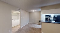 This single-floor residence offers a practical and well-appointed layout designed for comfortable everyday living. The open-concept living and dining area is finished with plush wall-to-wall carpeting and features a wood-burning fireplace with a shiplap surround, a ceiling fan with light fixture, and access to an exterior door that allows natural light to filter in. The kitchen is connected to the living space via a peninsula counter topped with dark granite, providing both a casual dining surface and a clear sightline into the main living area. The kitchen itself is equipped with black appliances including a refrigerator, microwave, and range, complemented by white cabinetry, dark granite countertops, a double stainless steel sink, and wood-look flooring. A dedicated in-unit laundry closet houses a full-size washer and dryer, adding significant convenience. The bedroom offers carpeted flooring, a ceiling fan, and a window with blinds for natural light and ventilation. The bathroom is finished with a dark granite vanity countertop, white cabinetry with ample storage, a framed mirror, and a lighted vanity fixture. Textured ceilings are consistent throughout the unit, and neutral wall tones create a cohesive and versatile interior palette.