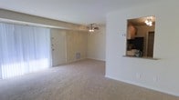 This single-story space presents an open and practical layout centered around a carpeted living area. The room is finished with neutral beige walls and white trim, creating a clean, understated aesthetic throughout. A ceiling fan with a light fixture is centrally mounted, providing both air circulation and ambient lighting to the space.

Natural light enters through a set of vertical blinds covering what appears to be a sliding glass door or large window along one wall, contributing to a bright and airy feel. A floor vent is visible along the baseboard, indicating a forced-air climate control system integrated into the unit.

A defining architectural feature is the pass-through opening in the partial wall that connects the living area to the kitchen. Through this opening, cabinetry, countertops, and kitchen appliances are visible, suggesting a functional kitchen space just beyond. This open connection between the two areas enhances the sense of flow and makes the overall layout feel more spacious.

The space is designed with simplicity and practicality in mind, offering a neutral palette and straightforward finishes that provide a versatile foundation for a variety of furnishing and décor styles.