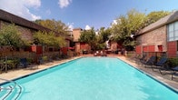 This community amenity space offers residents a range of thoughtfully designed areas for relaxation and socializing. At the heart of the outdoor area is a generously sized swimming pool with crystal-clear turquoise water, surrounded by a spacious pool deck lined with lounge chairs. Mature trees and brick-accented buildings frame the backdrop, creating a welcoming and shaded outdoor retreat.

Inside, the clubhouse lounge features warm hardwood-style flooring, crown molding, and a vaulted ceiling with ceiling fans and recessed lighting. The space is furnished with leather sofas arranged around a wall-mounted television, complemented by multiple round dining tables and chairs. Framed abstract artwork in muted tones adds a polished, contemporary touch to the sage-green walls.

The adjacent kitchenette is outfitted with dark espresso cabinetry, granite countertops, a stainless steel dishwasher, a water dispenser, toaster ovens, and microwaves. A striking mosaic tile backsplash serves as a bold focal point, illuminated by pendant lighting overhead.

A carpeted staircase leads to an upper-level business nook, offering a quieter area for focused work. Throughout the space, consistent finishes and carefully chosen décor create a cohesive, comfortable environment suited for both casual gatherings and everyday use.