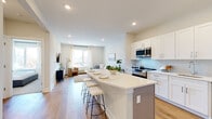 This single-floor residence is designed with a clean, contemporary aesthetic that emphasizes open space and natural light. The heart of the unit is an open-concept kitchen and living area, where warm hardwood-style flooring and recessed lighting create a welcoming atmosphere. The kitchen is well-appointed with white shaker cabinetry, quartz countertops, a light mosaic tile backsplash, and a full suite of stainless steel appliances, including a range, microwave, dishwasher, and refrigerator. A central island with seating provides a functional and social focal point, flowing naturally into the living area. The living space is bright and airy, benefiting from a large window that allows generous natural light and outdoor views. Neutral walls and light hardwood flooring carry throughout, maintaining a cohesive and calm palette. The two bedrooms are carpeted for comfort, each offering a tranquil retreat. The primary bedroom features a large window with outdoor views, while both rooms provide easy access to closet space. The full bathroom is finished with large-format tile flooring, a gray vanity with a white countertop, an oversized mirror, and a full soaking tub with shower combination. The overall ambiance is modern, airy, and thoughtfully designed.