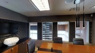This single-floor professional office space is designed to impress, combining functional design with a polished, contemporary aesthetic. The centerpiece of the space is a formal conference room outfitted with a warm-toned wood conference table surrounded by high-back leather executive chairs, creating an atmosphere of authority and professionalism. The walls are finished in deep, dark tones and adorned with textured geometric-patterned panels, adding visual depth and a refined character to the room. A striking ceiling-mounted light fixture with a luminous panel design provides bright, even illumination throughout the space. Interior windows with layered zebra-style roller blinds allow for adjustable privacy and light control, while also offering views into adjacent office areas, maintaining a sense of openness and connectivity within the workplace. Neutral-toned carpeting grounds the space, complementing the dark wall palette. The overall ambiance is one of focused professionalism — sophisticated, well-organized, and designed to facilitate productive meetings and executive discussions. Every detail, from the lighting to the furnishings, reflects a deliberate commitment to creating a high-functioning and visually cohesive professional environment.