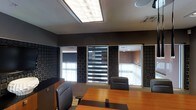 This single-floor professional office space is designed to impress, combining functional design with a polished, contemporary aesthetic. The centerpiece of the space is a formal conference room outfitted with a warm-toned wood conference table surrounded by high-back leather executive chairs, creating an atmosphere of authority and professionalism. The walls are finished in deep, dark tones and adorned with textured geometric-patterned panels, adding visual depth and a refined character to the room. A striking ceiling-mounted light fixture with a luminous panel design provides bright, even illumination throughout the space. Interior windows with layered zebra-style roller blinds allow for adjustable privacy and light control, while also offering views into adjacent office areas, maintaining a sense of openness and connectivity within the workplace. Neutral-toned carpeting grounds the space, complementing the dark wall palette. The overall ambiance is one of focused professionalism — sophisticated, well-organized, and designed to facilitate productive meetings and executive discussions. Every detail, from the lighting to the furnishings, reflects a deliberate commitment to creating a high-functioning and visually cohesive professional environment.