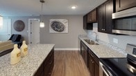This single-floor space offers a well-designed layout that balances functionality and style. The kitchen is a standout feature, showcasing rich dark-espresso cabinetry paired with light granite countertops that extend across both the perimeter counters and a central island. Stainless steel appliances, including a range, over-the-range microwave, and dishwasher, complement the modern aesthetic, while a pendant light adds a refined touch above the island. Warm-toned luxury vinyl plank flooring runs throughout the main living areas, creating a cohesive and easy-to-maintain surface.

The living area features a bold teal accent wall that provides a striking contrast to the neutral gray tones found throughout the rest of the space. Windows along the wall allow natural light to fill the room, enhancing its welcoming atmosphere.

The bedrooms are finished with plush carpeting and neutral taupe walls, contributing to a calm and restful environment. Each bedroom features windows with blinds that admit natural light while maintaining privacy.

The bathroom is finished with the same dark cabinetry and granite countertop found in the kitchen, ensuring a consistent design language. A walk-in shower with a white surround and wood-look flooring complete this clean, practical space.