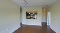 This single-floor residence offers a clean, contemporary design centered around an open-concept living area that feels bright and welcoming. Rich, dark hardwood flooring extends throughout the main living space, providing a warm contrast to the neutral, light-toned walls and white trim. A striking pass-through window connects the kitchen to the living area, creating a sense of openness while maintaining a defined cooking space. The kitchen is well-appointed with dark espresso cabinetry, stainless steel appliances including a range, microwave, and refrigerator, and a light-colored countertop that adds brightness to the space. Pendant lighting above the pass-through and under-cabinet illumination enhance the kitchen's functional ambiance. In the living area, a modern track lighting fixture provides adjustable overhead lighting, adding a stylish touch to the ceiling. A corridor visible from the main living space leads to additional rooms, with a paneled door offering privacy. The overall design aesthetic is modern and minimalist, combining practical layout choices with thoughtful finishing details that make the space feel polished and move-in ready.
