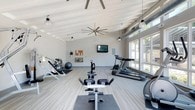 This single-floor fitness center offers a spacious and thoughtfully designed workout environment. The interior is defined by striking vaulted ceilings with exposed white-painted beams and visible ductwork, lending an industrial-chic character to the otherwise clean, modern aesthetic. Large windows along one wall flood the space with natural light while offering views of the surrounding greenery, creating an inviting and energizing atmosphere.

The flooring features light-toned, wood-look vinyl planking that adds warmth and is well-suited for high-traffic athletic use. Overhead, oversized ceiling fans ensure comfortable air circulation throughout the facility.

The equipment layout is well-organized and comprehensive. Cardio machines, including treadmills and an elliptical, are positioned along the windowed wall to take advantage of the natural light and outdoor views. A rowing machine station anchors the central floor space, while a dedicated free weights and functional training area — complete with a stability ball, weight rack, and incline bench — occupies the far end of the room. A wall-mounted television and bulletin board provide both entertainment and informational resources for users. Pendant and industrial-style hanging lights complement the overhead fans, ensuring the space remains bright and well-lit at any time of day.