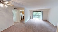This single-floor unit is thoughtfully laid out to maximize functionality across its living spaces. The main living area is generously sized, featuring neutral-toned wall-to-wall carpeting and crisp white walls that create a bright, open atmosphere. A large sliding glass door floods the space with natural light and provides direct access to a private balcony overlooking lush greenery, offering a pleasant outdoor view. A ceiling fan is installed in an adjoining room, adding to the unit's practical appeal.

The kitchen is well-equipped with warm-toned wood cabinetry offering ample upper and lower storage. White appliances, including a gas range and a refrigerator, are complemented by light countertops and a double-basin stainless steel sink positioned beneath a window that lets in natural light. The kitchen connects seamlessly to the living area through an open passageway.

A closet with louvered bifold doors is conveniently located near the living space. The bedroom areas are carpeted in neutral tones, with clean white walls and ample space. The bathroom features a full tub-shower combination with white tile surround, chrome fixtures and grab bars, a toilet, and a vanity with a light countertop, all finished with tile flooring for a clean, functional aesthetic.