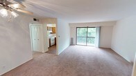 This single-floor unit is thoughtfully laid out to maximize functionality across its living spaces. The main living area is generously sized, featuring neutral-toned wall-to-wall carpeting and crisp white walls that create a bright, open atmosphere. A large sliding glass door floods the space with natural light and provides direct access to a private balcony overlooking lush greenery, offering a pleasant outdoor view. A ceiling fan is installed in an adjoining room, adding to the unit's practical appeal.

The kitchen is well-equipped with warm-toned wood cabinetry offering ample upper and lower storage. White appliances, including a gas range and a refrigerator, are complemented by light countertops and a double-basin stainless steel sink positioned beneath a window that lets in natural light. The kitchen connects seamlessly to the living area through an open passageway.

A closet with louvered bifold doors is conveniently located near the living space. The bedroom areas are carpeted in neutral tones, with clean white walls and ample space. The bathroom features a full tub-shower combination with white tile surround, chrome fixtures and grab bars, a toilet, and a vanity with a light countertop, all finished with tile flooring for a clean, functional aesthetic.
