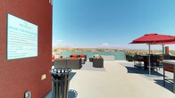 This rooftop terrace is a thoughtfully designed outdoor amenity space that takes full advantage of its elevated position, offering sweeping panoramic views of a surrounding desert landscape, distant mountain ranges, a nearby lake or reservoir, and a city skyline on the horizon. The terrace is finished with a smooth, light-toned concrete deck and is organized into several distinct functional zones. A central lounge area features multiple wicker sectional sofas and armchairs with bold orange cushions, arranged around a built-in rectangular gas fire pit table — ideal for gathering during cooler evenings. Adjacent to this is a sunbathing zone equipped with modern chaise loungers upholstered in teal, positioned to maximize sun exposure and views. A dedicated bar and dining area includes high-top tables and bar stools shaded by large red market umbrellas, alongside a built-in counter with bar-height seating. An outdoor kitchen station, sheltered beneath a retractable pergola-style cover, houses a stainless steel built-in grill and is outfitted with speakers and counter space. A glass railing system along the perimeter ensures unobstructed sightlines across the cityscape. The building's bold red and teal exterior accents provide a vibrant architectural backdrop throughout the space.