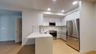 Located at 1669 Lincoln Ct in Santa Monica, California, this thoughtfully designed single-floor residence showcases a clean, contemporary aesthetic throughout. The kitchen serves as the heart of the space, boasting high-gloss white cabinetry that extends to the ceiling, a striking marble-veined quartz backsplash, and matching quartz countertops. A peninsula with an integrated sink provides additional prep space and casual seating potential. Stainless steel appliances, including a range with oven, over-the-range microwave, and a full-size refrigerator, complete the well-appointed cooking area. Light bamboo-style hardwood flooring runs throughout the main living areas, adding warmth and continuity. The bedrooms are bright and airy, with large windows framing views of the surrounding greenery and allowing generous natural light to fill the rooms. One bedroom features a ceiling fan and a built-in closet with shelving for organized storage. The bathroom is finished in a crisp, all-white palette with geometric marble-patterned tile flooring, a soaking tub and shower combination, and a modern vanity topped with a white stone surface and an illuminated mirror, lending a spa-like feel. Recessed lighting throughout enhances the clean lines and modern sensibility of this Santa Monica residence.