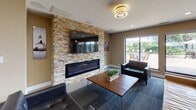 Located at 1647 Westminster Dr in Naperville, IL, this single-floor TGM Communities clubhouse is a well-appointed amenity space designed to serve as a social and functional hub. The lounge area immediately draws attention with its striking floor-to-ceiling stacked stone accent wall, which frames a sleek linear electric fireplace and a large wall-mounted television. Comfortable leather seating and a mid-century modern coffee table are arranged on patterned carpet, creating an inviting gathering space. Sliding glass doors open to views of an outdoor pool and patio area, allowing natural light to fill the room. The adjacent dining and café area features a bold red accent wall adorned with vibrant Chicago cityscape artwork, adding energy and color to the space. A built-in coffee and beverage station with dark granite countertops and charcoal cabinetry serves the dining tables, which are lined with modern white chairs on light-toned tile flooring. A dedicated business center runs along one wall, equipped with multiple computer workstations and high bar seating, providing a practical workspace within the communal setting. Together, these areas offer a seamless blend of comfort, functionality, and contemporary design throughout the space.
