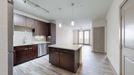 This one-bedroom, one-bathroom unit in San Antonio, TX offers a efficiently designed single-floor layout with a cohesive blend of modern finishes and neutral tones. The open-concept kitchen and living area serves as the heart of the space, anchored by light wood-style plank flooring that extends throughout. The kitchen is well-equipped with rich dark-walnut cabinetry reaching to the ceiling, granite countertops, a glass tile backsplash, stainless steel appliances including a range, microwave, dishwasher, and refrigerator, and a central island with additional counter space. Pendant lighting and under-cabinet lighting add functional illumination. The adjacent living area is generously proportioned, featuring a ceiling fan and track lighting. A hallway connects the main living space to the private bedroom, which features plush carpet flooring, a ceiling fan, and a window offering natural light. The bedroom is served by a spacious walk-in closet outfitted with multiple hanging rods and built-in shelving on all sides. The full bathroom includes a dark-wood vanity with granite countertop, tile flooring, and access to both a soaking tub and a separate tiled shower.
