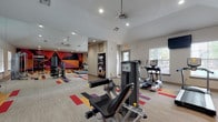 This fitness center is a thoughtfully designed workout facility that combines energetic aesthetics with a comprehensive range of equipment. The space is characterized by vaulted ceilings that lend an open, airy feel, complemented by recessed lighting and ceiling fans that maintain comfort during workouts. Large windows along the perimeter allow natural light to filter in, providing views of the surrounding outdoor areas.

The flooring features a neutral base with bold, geometric carpet tile accents in red, orange, purple, and black — a design motif echoed by the striking large-scale geometric mural that anchors one end of the room, adding a dynamic and motivating atmosphere.

Equipment is well-distributed throughout the space. Cardio offerings include treadmills, ellipticals, and a stair climber, all positioned near the windows. A strength training section includes selectorized cable machines and a leg press, while a dedicated free weight area provides tiered dumbbell racks and open floor space. A wall-mounted functional training rig stocks kettlebells, medicine balls, TRX straps, and foam rollers. Floor-to-ceiling mirrors visually expand the space and aid in form monitoring. Wall-mounted televisions are positioned for easy viewing. A snack vending machine is conveniently accessible near the entrance.